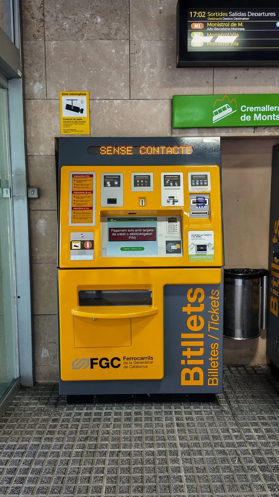 Yellow and grey FGC ticket machine at the Montserrat train station with a digital departures board above.